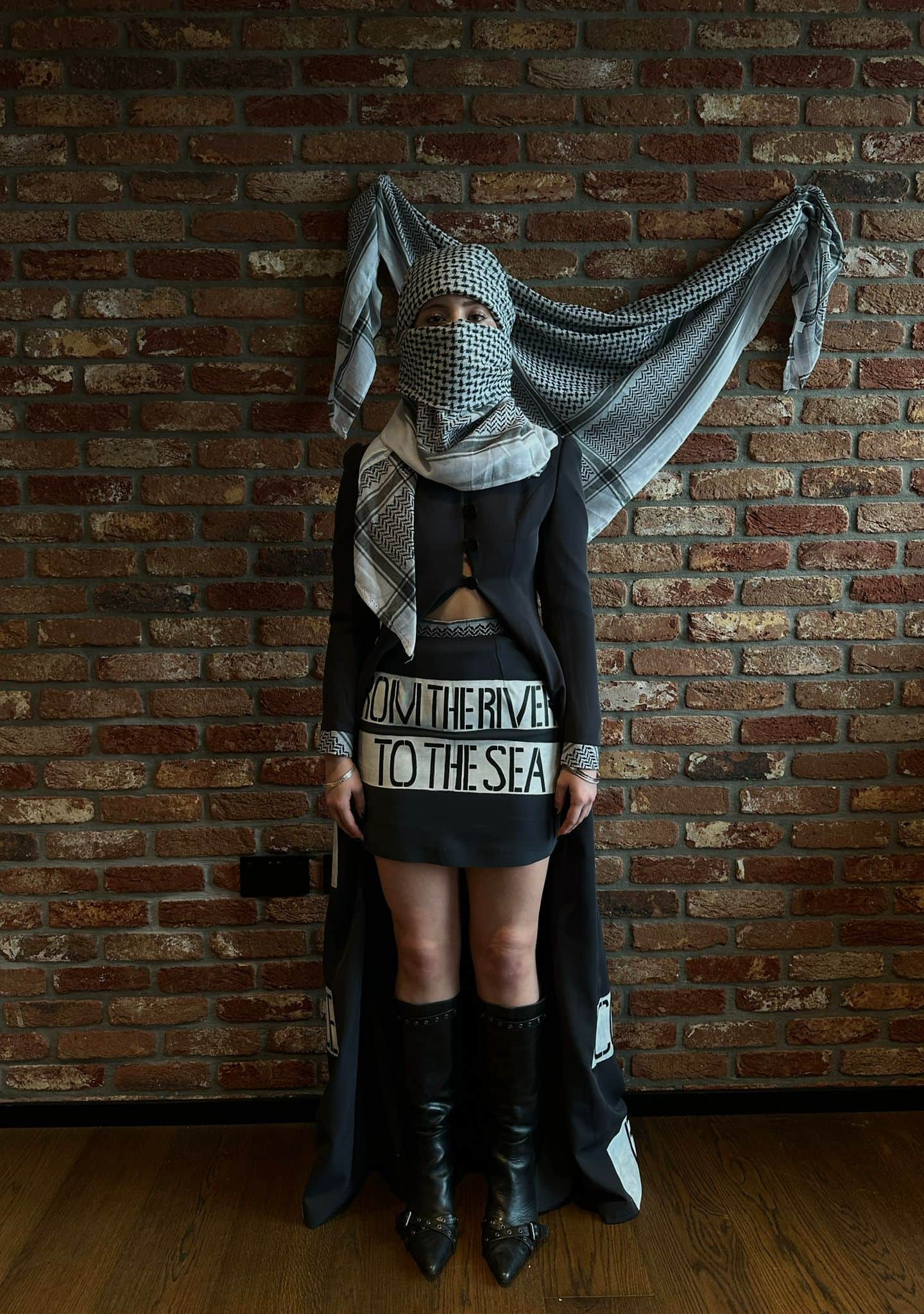 Atelier Qeblawi Coat of Resistance — short view, model in a charcoal grey thick-crepe coat with kufiyeh head wrap, knee-high boots, and a waist sash hand-painted 'From the river to the sea', against a brick wall with the kufiyeh draped behind.