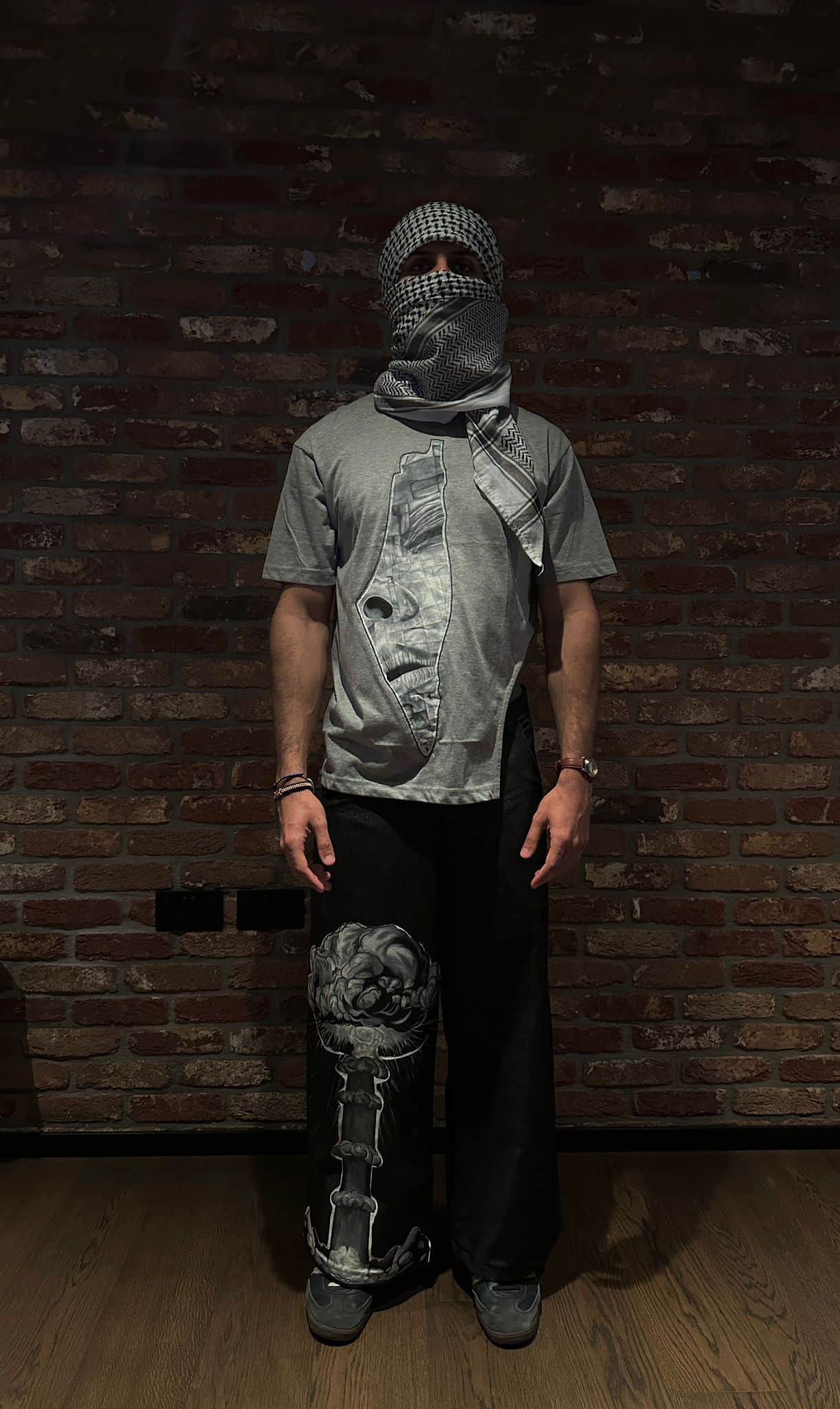 Atelier Qeblawi Painted Streetwear — front view, model in a grey cotton-jersey t-shirt with a hand-painted kufiyeh-veiled portrait and wide-leg dark denim jeans with a painted knee detail, kufiyeh head and face wrap, against a brick wall.
