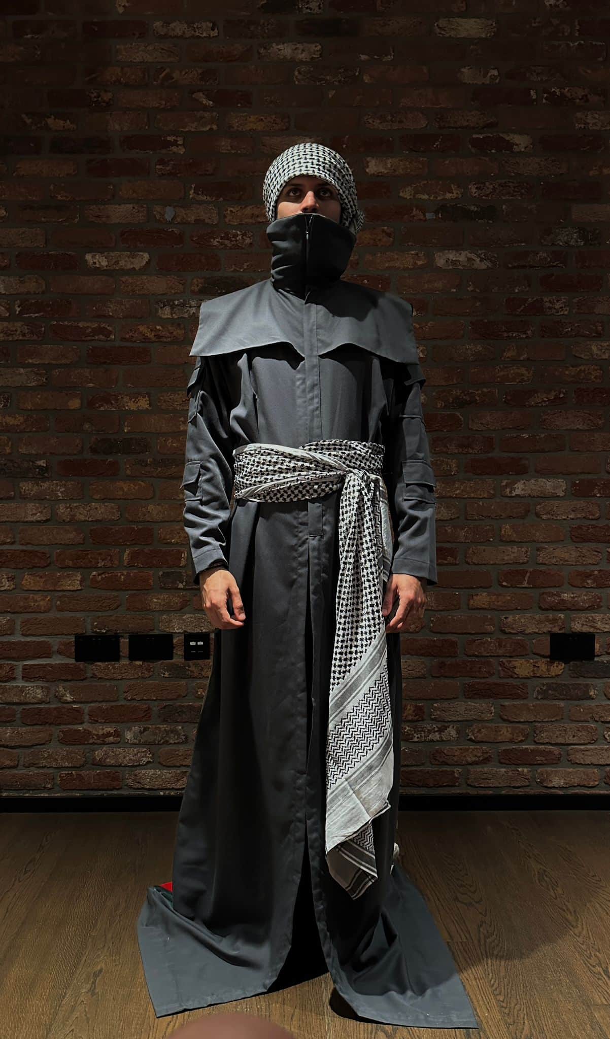 Atelier Qeblawi Witness Coat — front view, model in a floor-length grey gabardine coat with shoulder cape, kufiyeh head wrap and kufiyeh waist sash, against a brick wall.