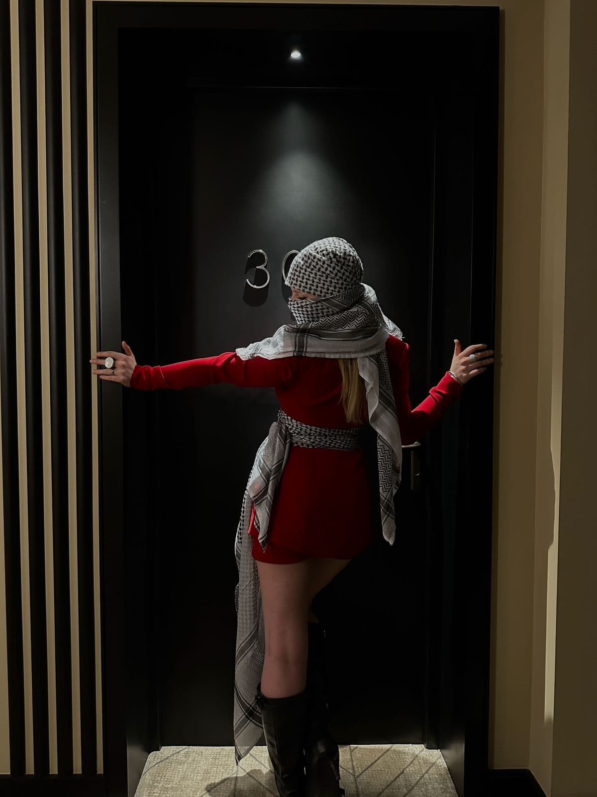 Atelier Qeblawi Crimson Tailoring — front view, structured red blazer-dress with kufiyeh head-wrap and waist sash, photographed in a darkened doorway.