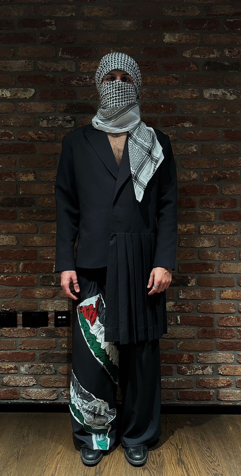 Atelier Qeblawi Painted Suit — front view, tailored black two-piece worn with kufiyeh head-wrap, against an exposed brick wall.