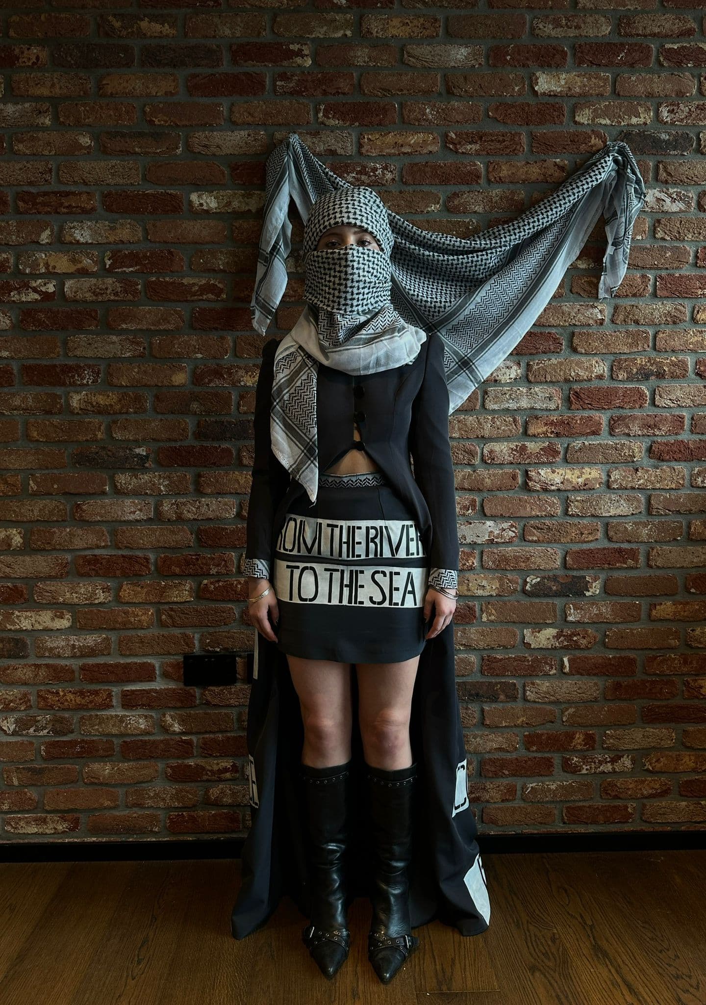 Atelier Qeblawi Coat of Resistance — short view, model in a charcoal grey thick-crepe coat with kufiyeh head wrap, knee-high boots, and a waist sash hand-painted 'From the river to the sea', against a brick wall with the kufiyeh draped behind.
