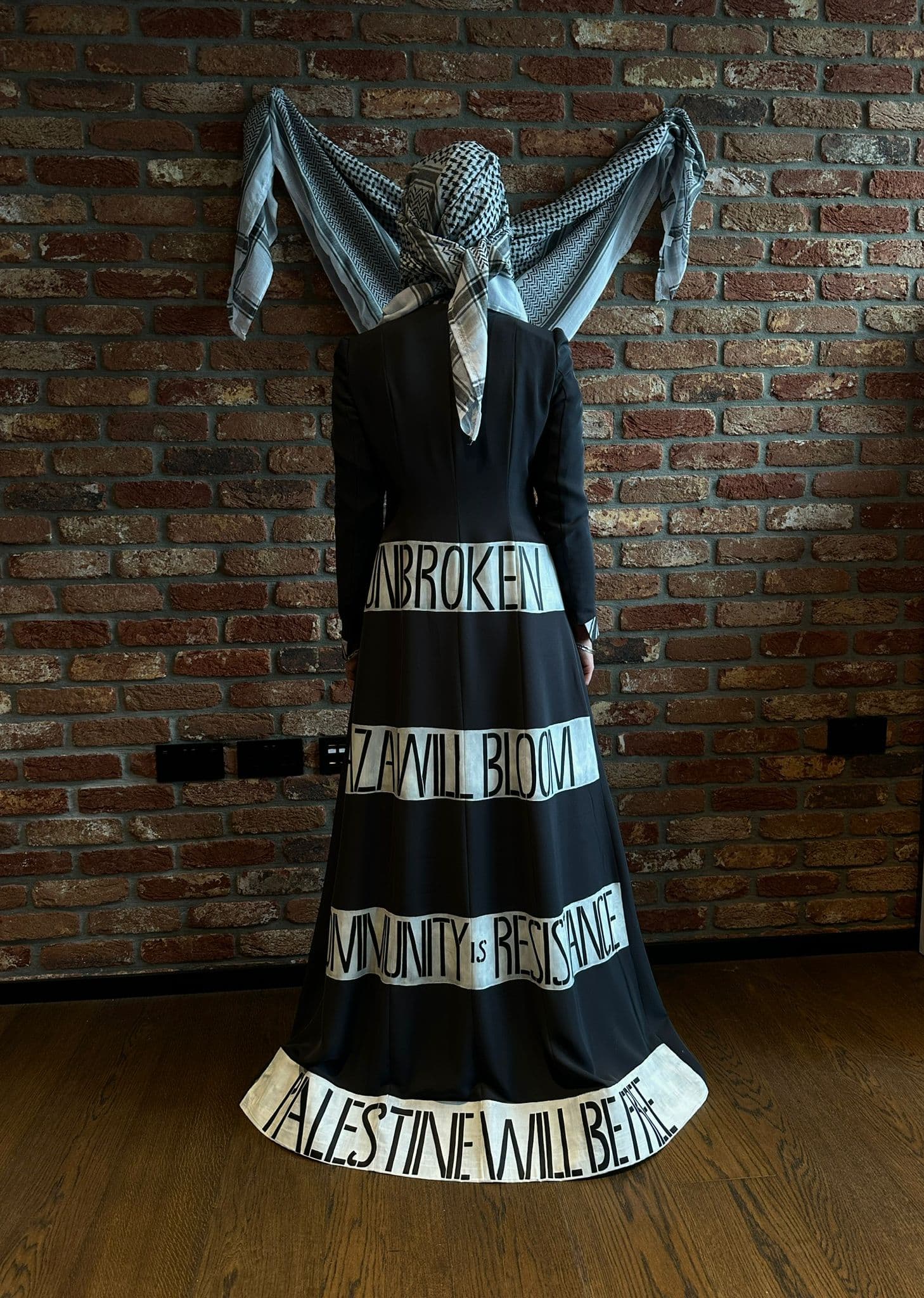 Atelier Qeblawi Coat of Resistance — long view, model in a floor-length charcoal grey coat with hand-painted text descending the body: 'Unbroken', 'Community is resistance', 'Palestine will be free'.