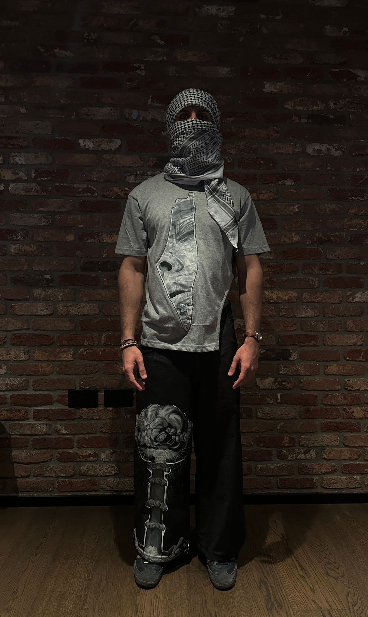 Atelier Qeblawi Painted Streetwear — front view, model in a grey cotton-jersey t-shirt with a hand-painted kufiyeh-veiled portrait and wide-leg dark denim jeans with a painted knee detail, kufiyeh head and face wrap, against a brick wall.