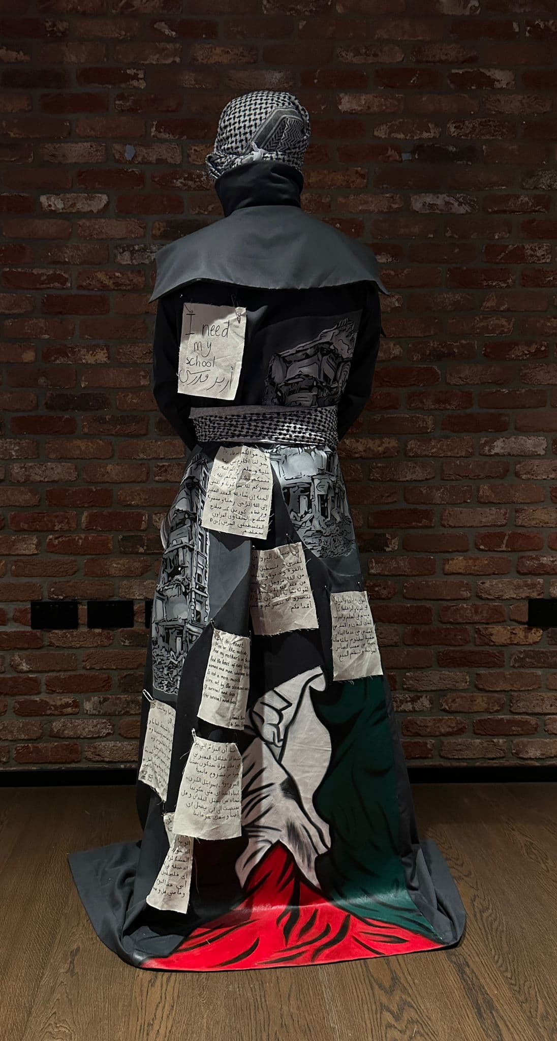 Atelier Qeblawi Witness Coat — back view, hand-painted scene of destruction and a Palestinian flag in red, white, green and black on the body of the coat, pinned with handwritten paper testimonies.