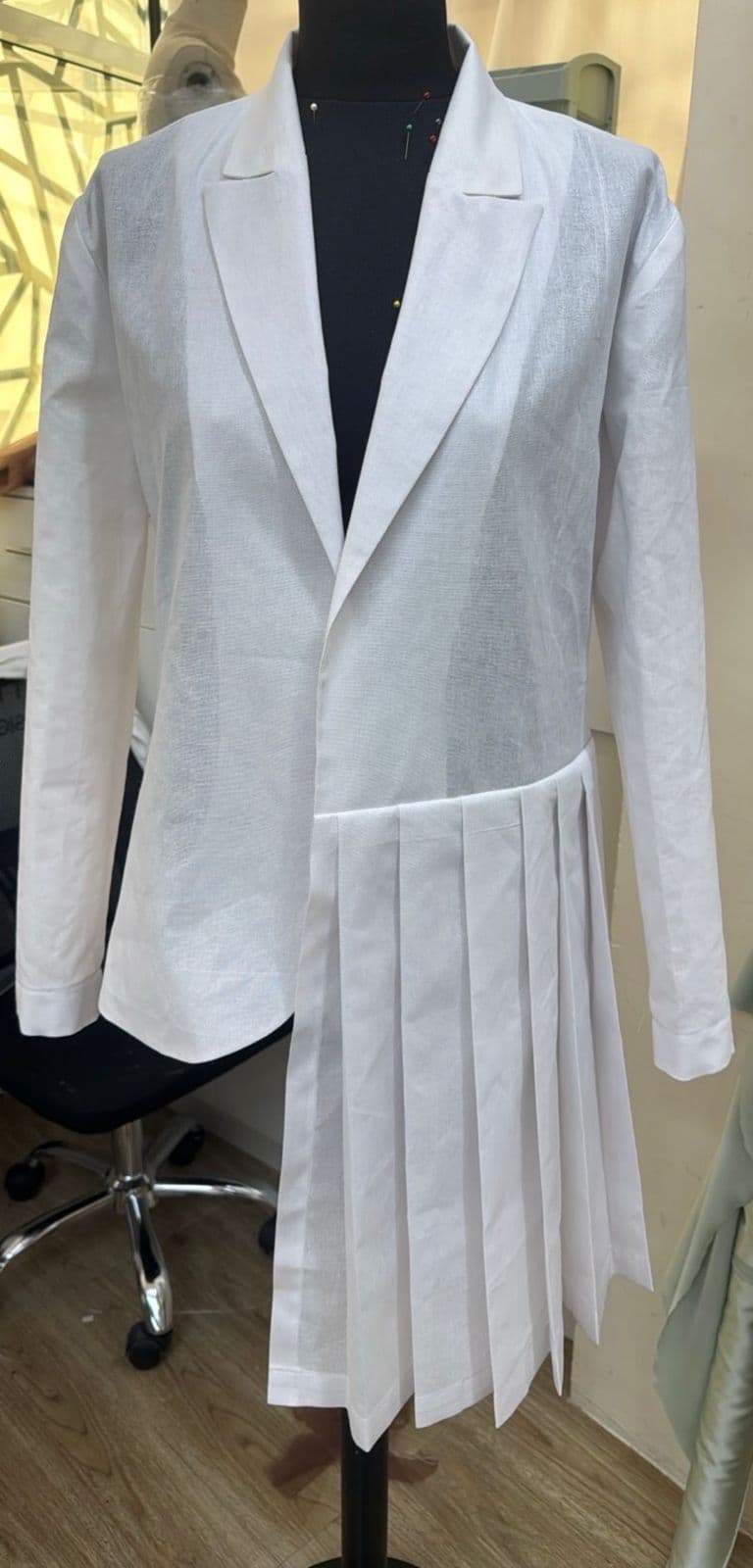 A white tailored blazer-dress with asymmetric pleated half-skirt panel pinned on a dressmaker's mannequin.
